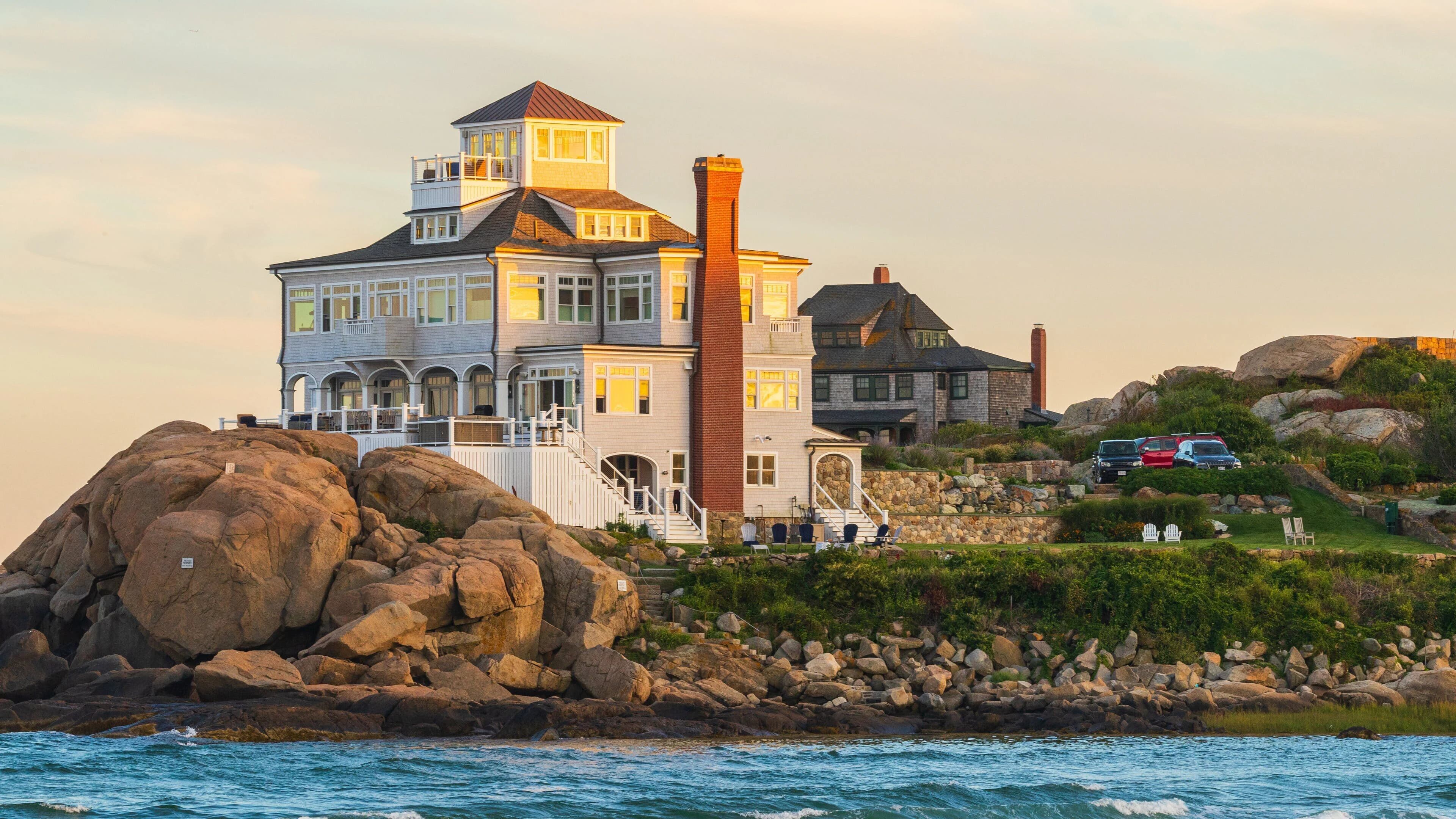 A Cape Ann waterfront house with chimney and lit windows at golden hour
